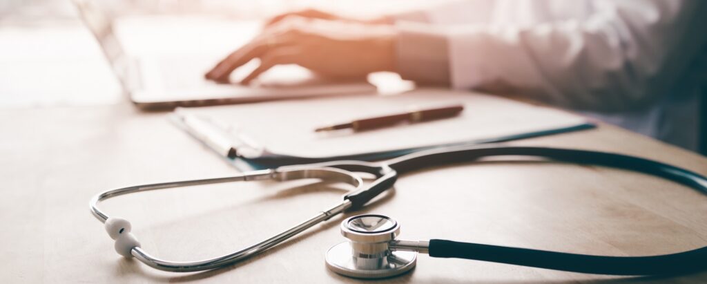 Stetoskop w tle lekarza pracującego na laptopie w gabinecie, symbol połączenia technologii i medycyny.