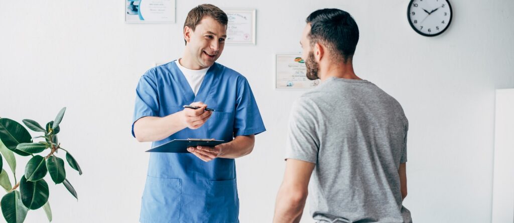Uśmiechnięty fizjoterapeuta - ubezpieczony w INTER Polska - z diagnozą w ręku wyjaśnia terapię pacjentowi w szpitalu.
