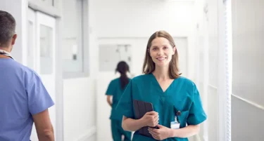 Pielęgniarka stoi na korytarzu szpitala i uśmiecha się do obiektywu; jest objęta ubezpieczeniem zdrowotnym dla medyków od INTER Polska.