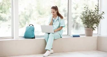 Studentka medycyny siedzi na korytarzu uniwersytetu z laptopem i kupuje ubezpieczenie zdrowotne INTER Polska online.