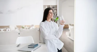 Lekarka stoi w salonie ze szklanką wody, zadowolona z wykupionego ubezpieczenia mieszkania i oferty INTER Polska.